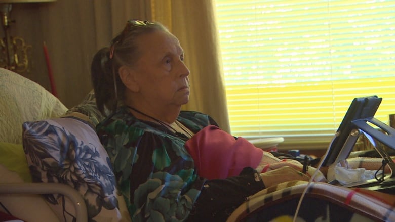 A woman lays in her hospital bed at home covered by blankets. A tablet sits on her lap. 