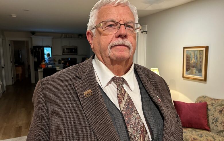 An older man with a large mustache and glasses, wearing a brown jacket, cardigan vest and shirt and tie, stands in a living room.  