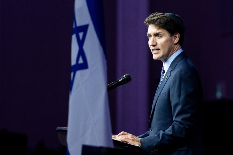 A man speaks into a microphone. An Israeli flag is drapped in the background.