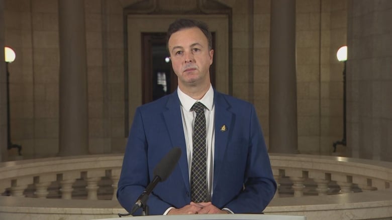 A man in a blue suit, white shirt and checkered tie looks forward from his position behind a podium.