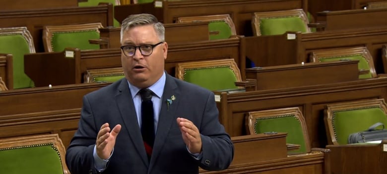 Man in blue suit and glasses standing up, behind him are wooden desks and chairs padded with green.