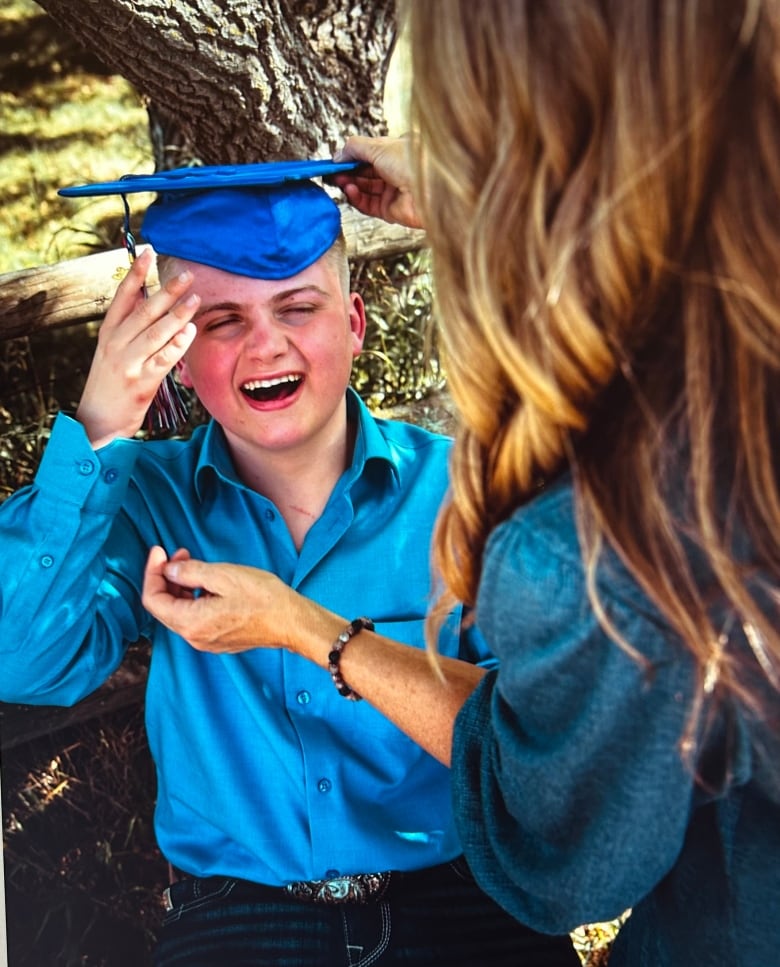 A smiling boy in blue tries on a graduation hat that a woman in the foreground adjusts over his head.