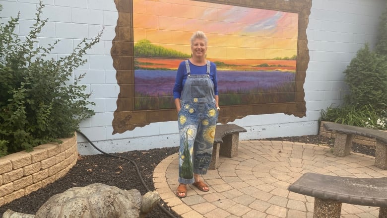 A white-haired woman in colourfully painted overalls stands in front of a mural depicting a skyscape.