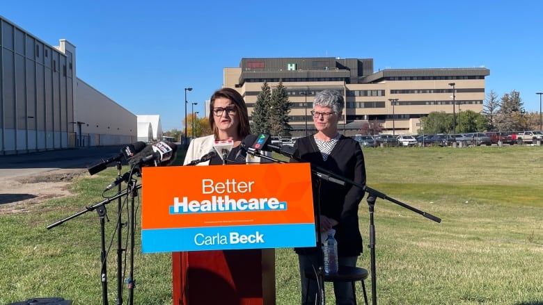 Two women stand at a podium. One woman is wearing a white shirt and a black sweater. To that woman's left and in front of her is a woman wearing a white jacket. Behind the podium is a grass field and then a hospital. 