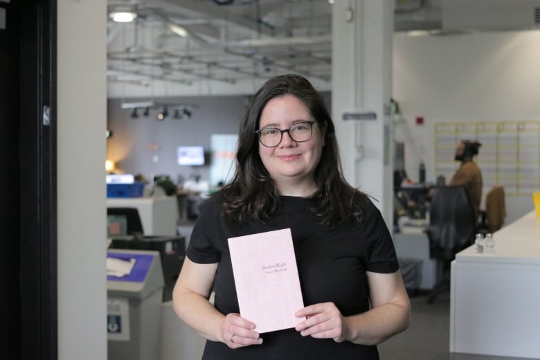 A person with long hair wearing glasses smiles as she holds a book. She's in an office.