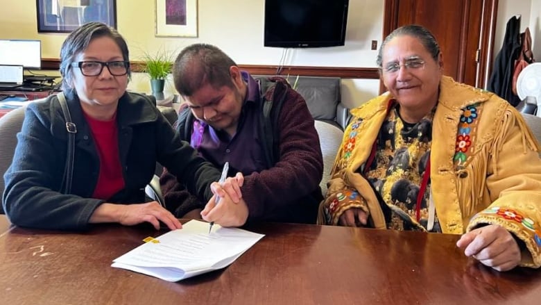 Three people smile at a table as one signs a document.