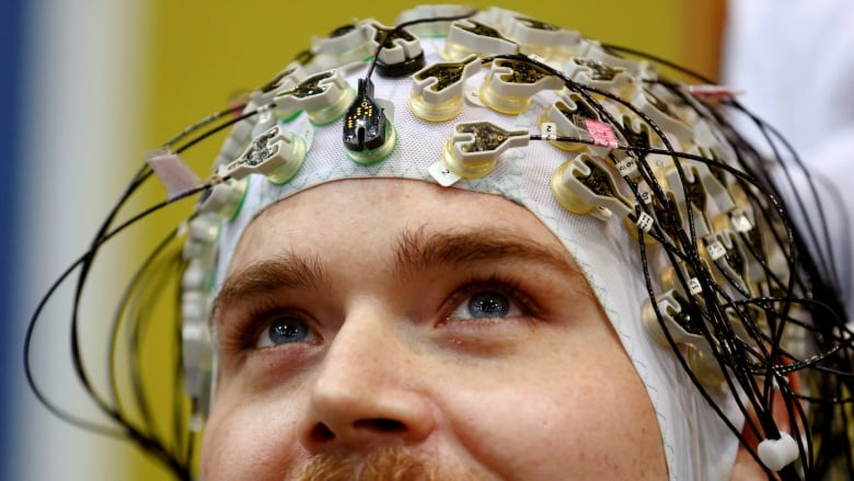 A mustachioed man smiles and looks up while wearing a skullcap covered in wires attached to electrodes