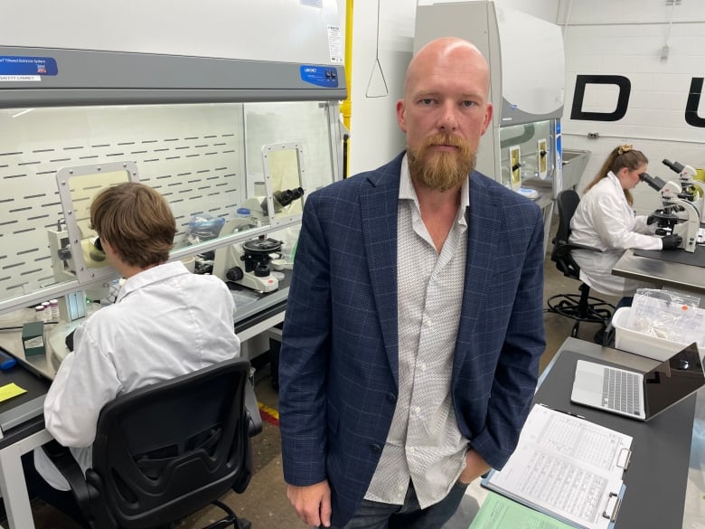 A man wearing a blue suit jacket is standing in a laboratory where two technologists wearing white lab coats are seated and looking into microscopes.