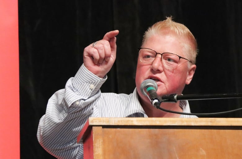 A woman with short blonde hair and glasses wearing a pinstriped white shirt points her finger and speaks into a microphone.