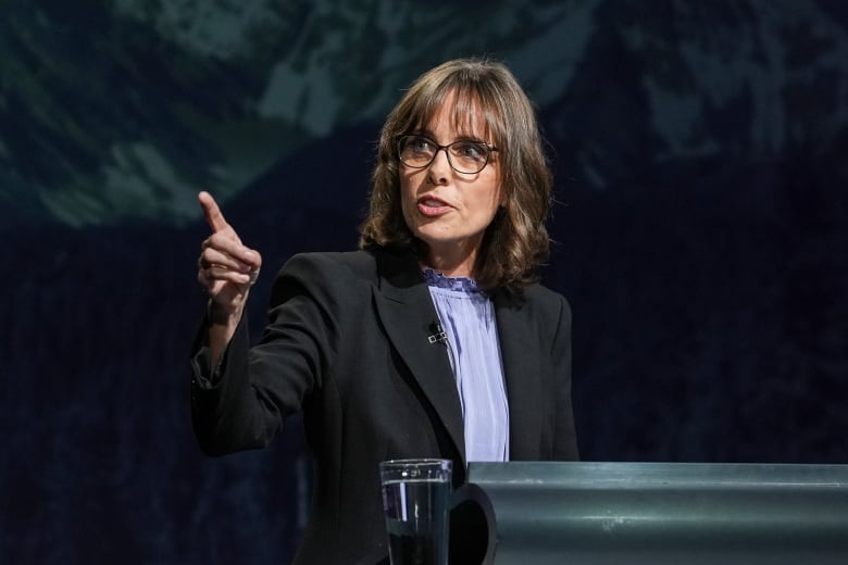 A woman points to her right while in front of a mountain backdrop.