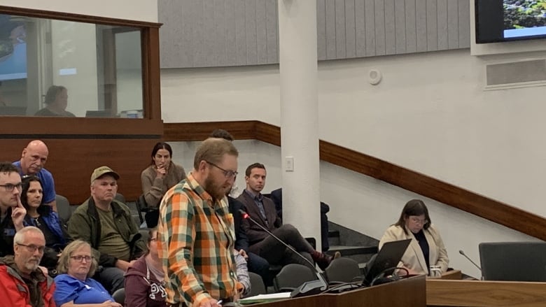 Man wearing plaid shirt speaking at a podium with a group of people behind him.