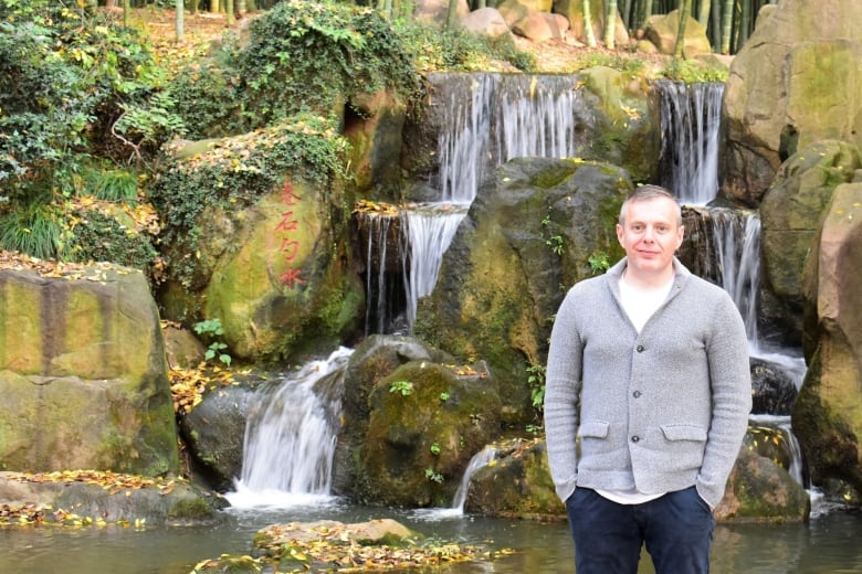 A man in a grey sweater is shown standing next to a small waterfall.