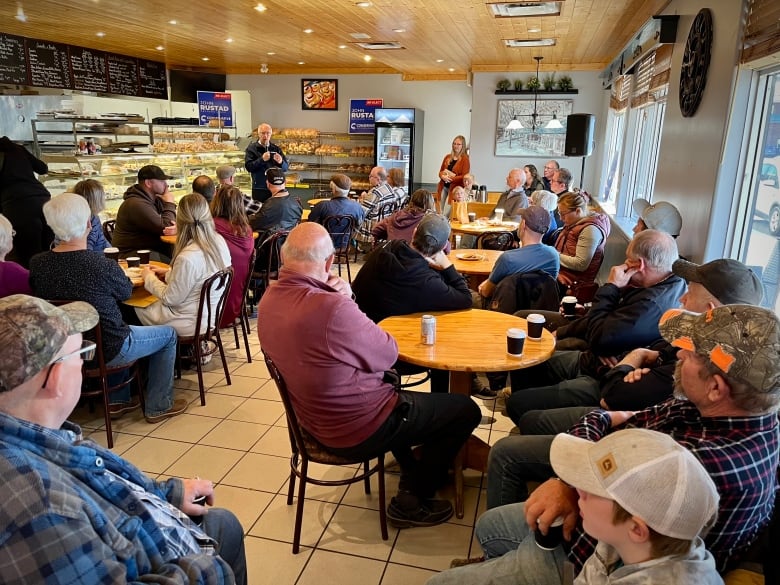 A man speaks to a crowd seated in a donut shop. 