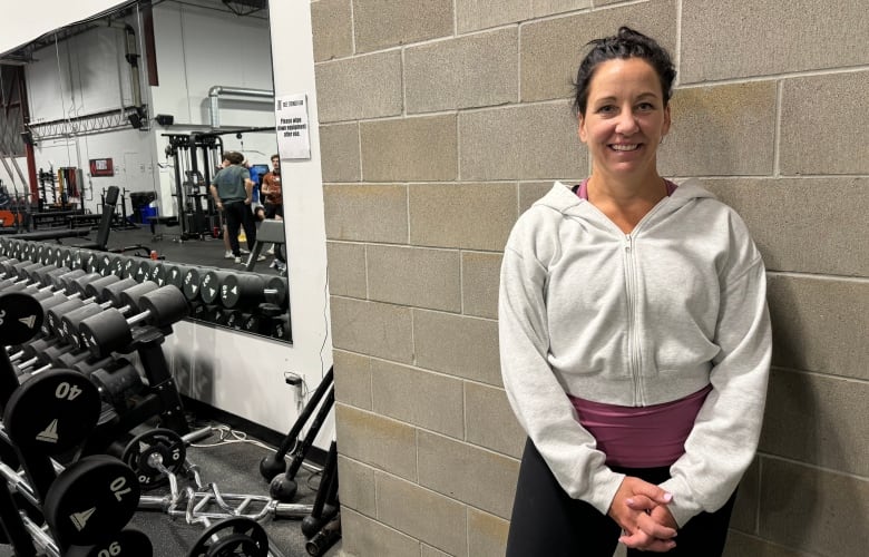 A smiling woman in a white hoodie and yoga pants can be seen next to a mirror and dumbbells at a gym.