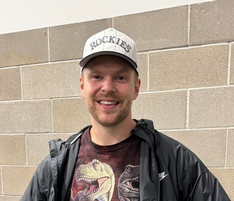 A bearded man in a jacket and ball cap stands with his arms behind his back.