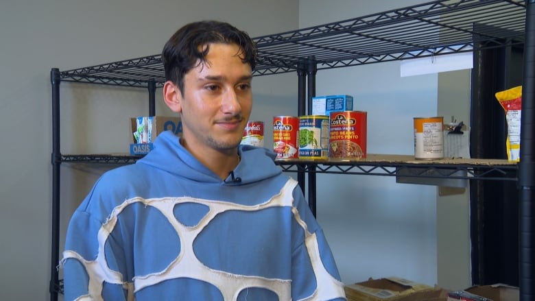 A man with short brown hair wears a blue and white hoodie. He's standing in front of a set of black shelves with a few cans of food on it. 