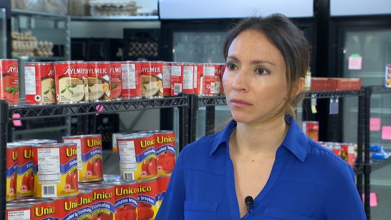 A woman wears her brown hair pulled back in a ponytail. She is wearing a royal blue blouse and stands in front of a shelf stocked with cans of soup and tomato sauce. 