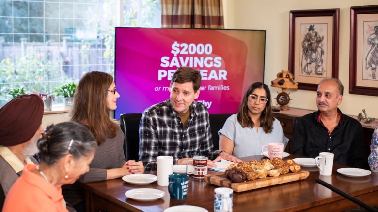 A group of people sit around a table in discussions with a man in a chequered shirt with an LCD TV behind him portraying the words, $2,000 Savings per Year.