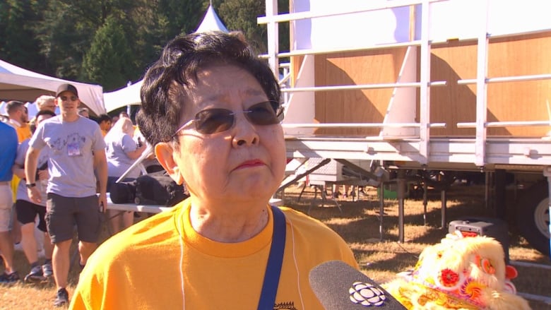 An East Asian woman in a yellow shirt speaks outdoors.