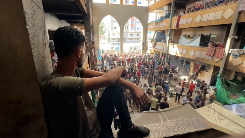 A man sits on the ledge of a balcony and overlooks people below.