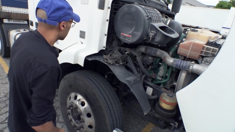 A man in a blue hat inspects the engine of a big rig truck.