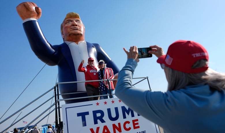 In the foreground, which is out of focus, a person holds up a phone camera to take a picture. A couple stand on top of a structure that says TRUMP VANCE on it, with a giant blow-up of Donald Trump behind them. 