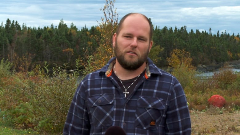 A man wearing a blue flannel shirt stands in a grassy area by a lake.