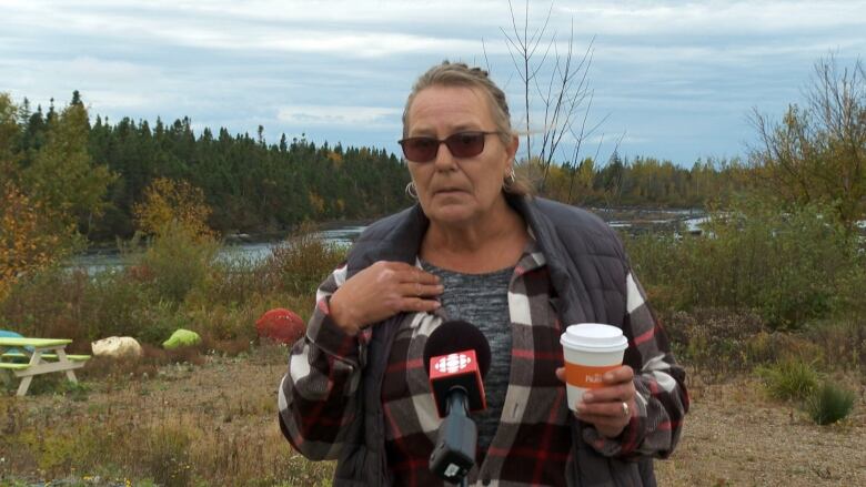 A woman wearing a vest speaks into a microphone while holding a paper cup of coffee.