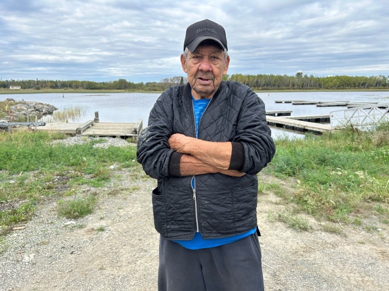 A man wearing a black hat, blue shirt and black jacket is pictured in front of docks at the mouth of a river.