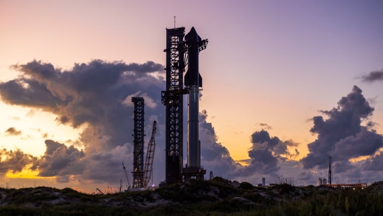 A rocket is seen sitting on a launch pad with an orange and pink sky with some clouds.