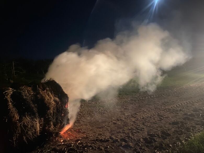 A hay bale burns at night, producing smoke that ripples across a farm field.