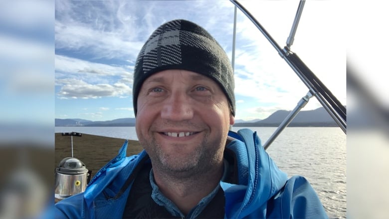 A man wearing a black and grey checkered toque and blue coat smiling at the camera. 