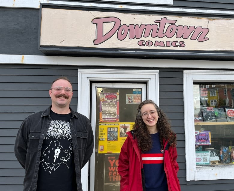 A person in black shirt standing next to woman with long brown hair in a red jacket. Both are smiling and behind them is a doorway and a sign that says 