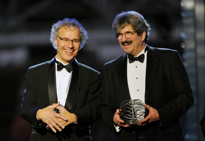 Two men wearing tuxedos smile past the camera. They are holding round awards.