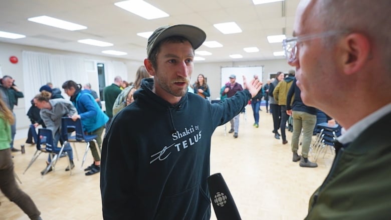 A man in a black hoody with a baseball hat on gestures as he answers a question from a reporter in a hall full of people.