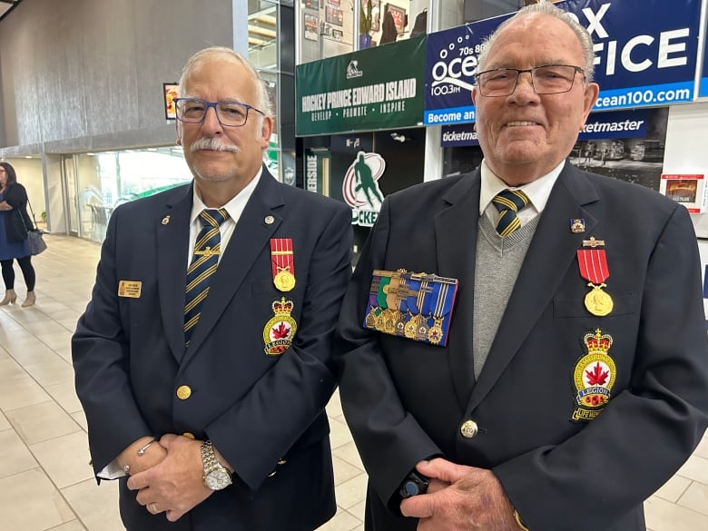 Two older men wearing air force uniforms with several medals stand next to each other in the lobby of a sports arena. 