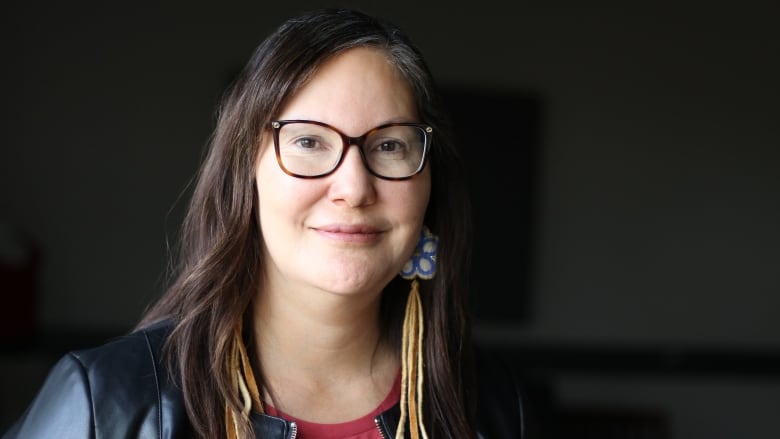 A woman with long brown hair and black square frame glasses is photographed smiling. 