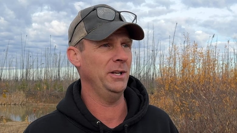 Middle aged man, photographed from chest up. Wearing baseball cap with glasses. Trees and shrubs in the background. 