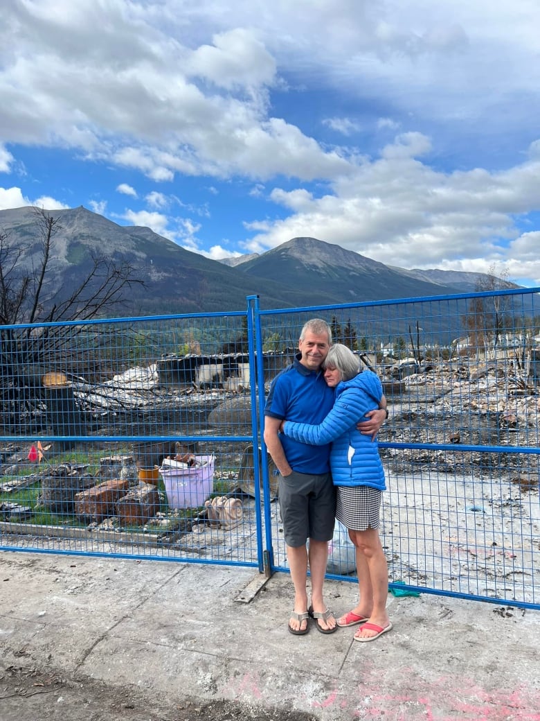 A man and woman stand hugging each other in front of a tall construction fence. Behind them, their home is completely burned down. All that's left are grey and black charred pieces of foundation, a few Tupperware containers and some gardening supplies.