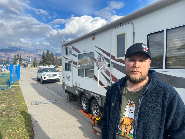 A man in a ball cap stands in front of a trailer, parked on a street in Jasper, Alta. There's rubble in the background of houses that were destroyed in July's wildfire.