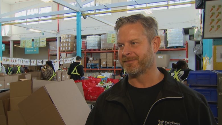 A man stands in a warehouse with volunteers filling boxes behind him. 