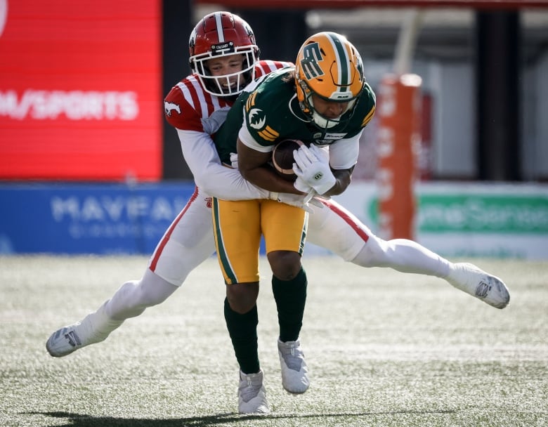 A football player in a green-and-yellow uniform is cradling the ball, while an opponent in a red-and-white uniform tackles him from behind.