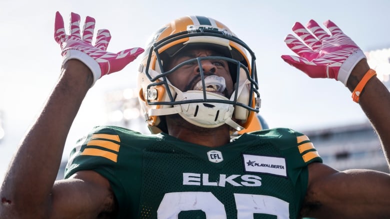 A Black football player wearing a green-and-yellow uniform, and red-and-white gloves, is celebrating on the field.