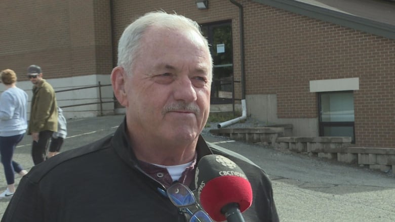 A man with thinning white hair and a mustache stands outside a brick building, speaking into a microphone.