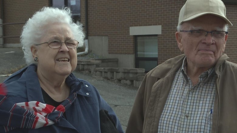 One elderly woman and one elderly man stand outside in front of a brick building.