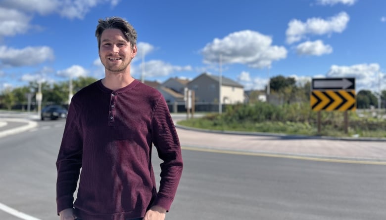 A white man in a burgundy sweater smiles in front of a roundabout.