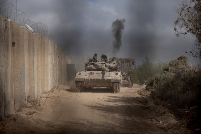 Tanks navigate a dirt road near a wall topped with barbed wire.