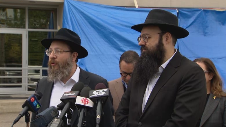 Two rabbis speak in front of media microphones outside, with a blue tarp behind them