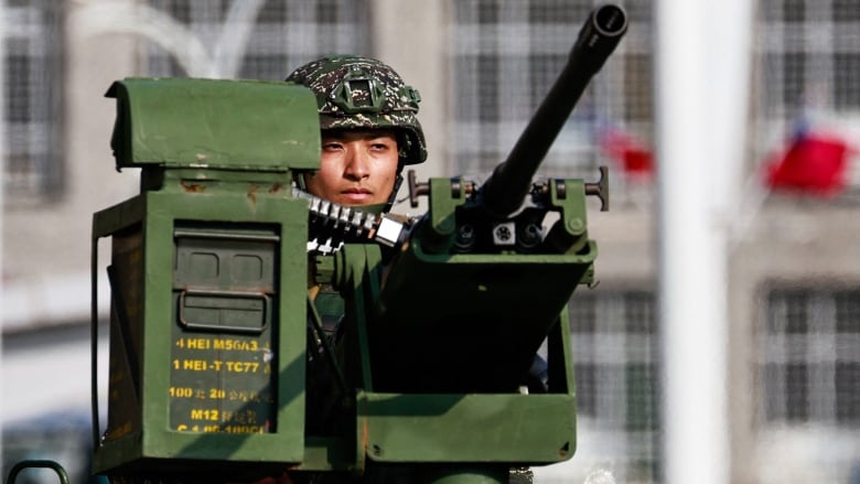 A helmeted man's head is visible behind a large gun in military green. Buildings are out of focus in the background.