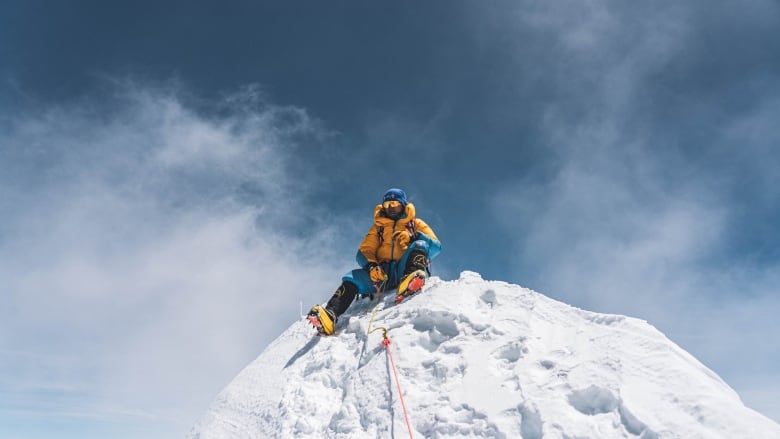 A climber in full orange and blue gear sits atop a snowy peak with only sky and clouds above him.